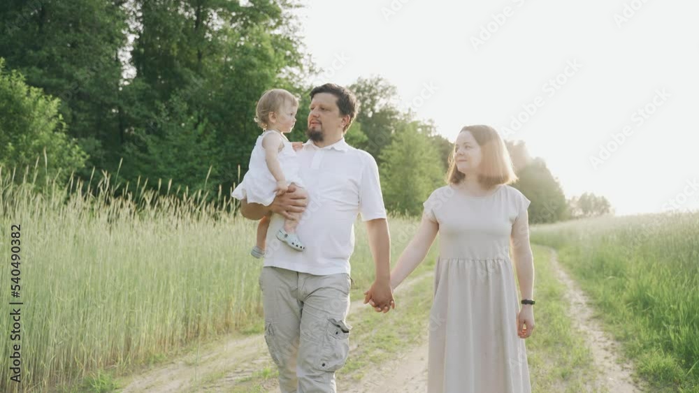 Happy parents hold a child in their arms and walk along the road along the field in the rays of the sunset field. Family and children concept. Happy parents hold a child in their arms and walk along the road along the field in the rays of the sunset field. Family and children concept.
