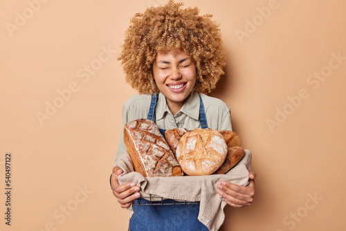 Joyful curly haired female baker holds basket of freshly baked bread smiles happily glad to finish work shows various types of bakery wears shirt and denim sarafan isolated over beige background photo