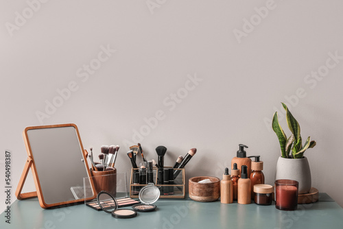 Different cosmetics with mirror and houseplant on table near light wall