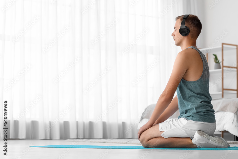 Fototapeta premium Young man with headphones meditating on mat in bedroom