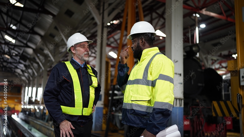 Engineers in discussion in machine maintenance in factory. Industrial Engineers with Blueprint against trains and machinaries industry background. Studying Blueprint with Superior Engineers in discussion in machine maintenance in factory. Industrial Engineers with Blueprint against trains and machinaries industry background. Studying Blueprint with Superior