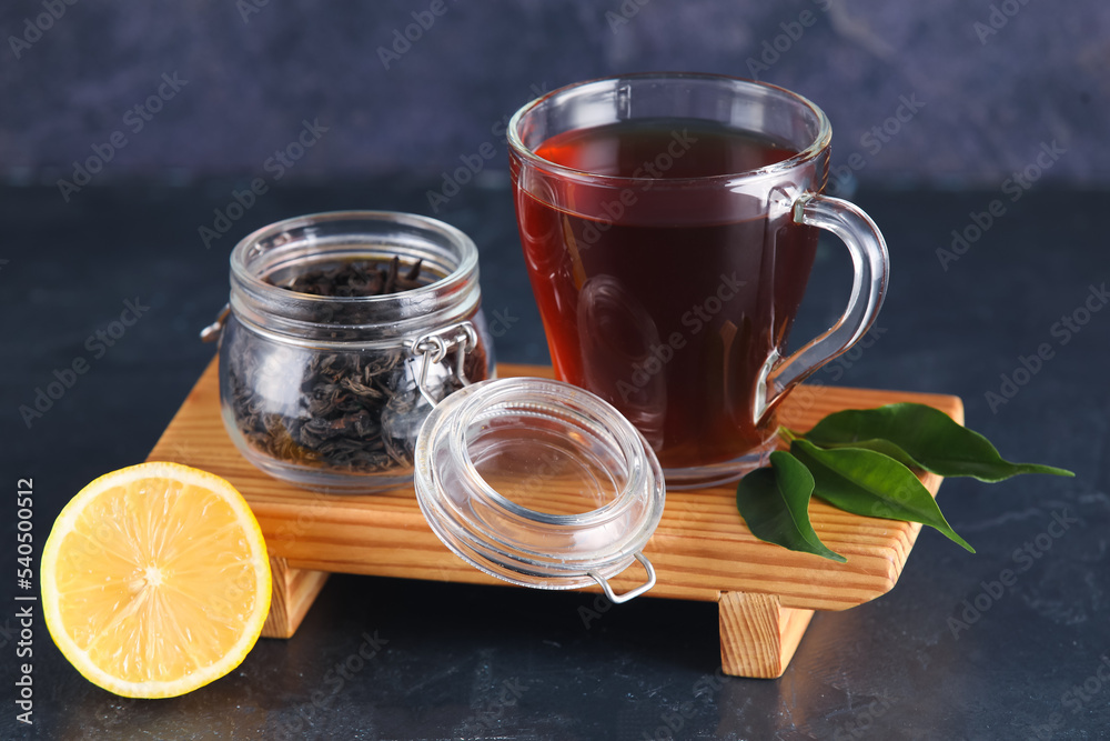 Wooden board with glass cup of black tea and dry leaves on dark background Wooden board with glass cup of black tea and dry leaves on dark background