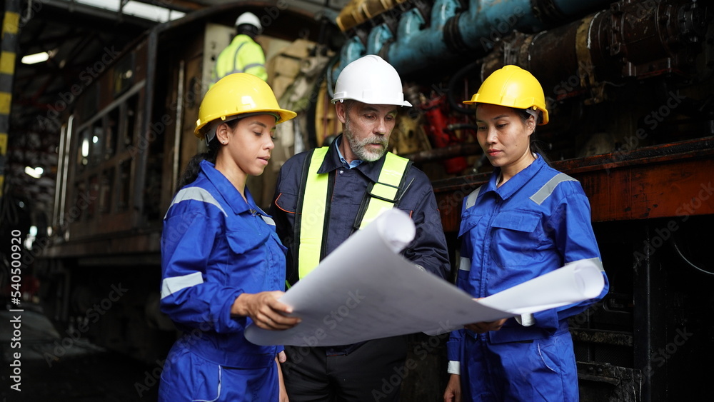 Engineers in discussion in machine maintenance in factory. Group of Industrial Engineers with Blueprint against trains and machinaries industry background. Engineers in discussion in machine maintenance in factory. Group of Industrial Engineers with Blueprint against trains and machinaries industry background.