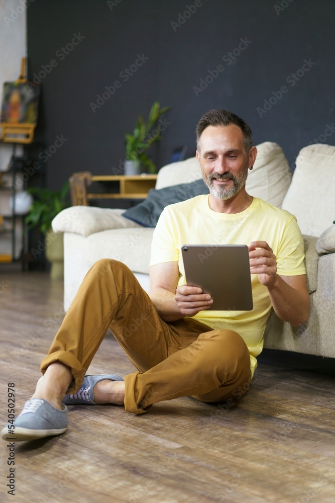 Student mature man using tablet pc, browsing internet application on digital tablet, sitting on the floor over flat interior and lean on coach