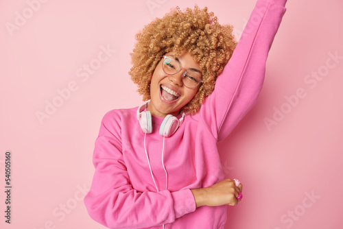 Horizontal shot of optimistic woman dances carefree foolishes around has fun listens favorite music wears pullover headphones around neck keeps arm raised smiles cheerfully isolated over rosy wall