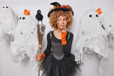 Serious thoughtful curly haired witch looks attentively aside wears black hat with autumn leaves and dress holds broom surrounded by spooky creatures celebrates Halloween on party. Trick or treat