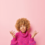 Indifferent clueless woman with curly hair spread palms and feels doubtful focused overhead stands questioned against pink background wears knitted jumper copy space above cannot make decision