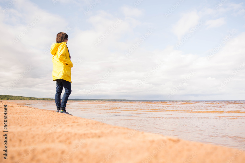 Faceless woman in yellow raincoat standing on beach and looking at sea 