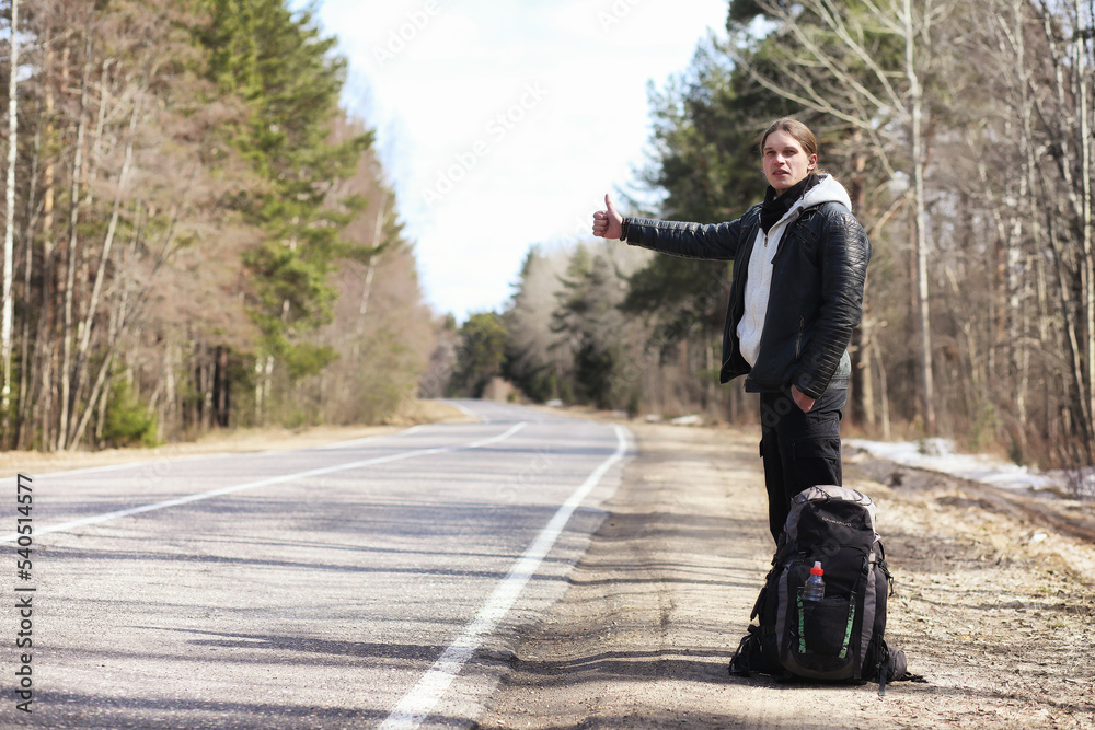 A young man is hitchhiking around the country. The man is trying to catch a passing car for traveling. The man with the backpack went hitchhiking to the south.
