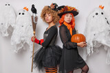Horizontal shot of attentive women witches stand backs with broom and pumpkin await for Halloween wear black dresses stand against spooky creatures prepare for celebration. Witchcraft concept