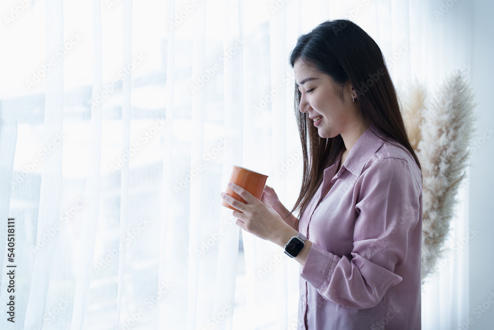 A portrait of a young Asian woman in pajamas smiling happily by a window with curtains in the morning sun while drinking coffee A portrait of a young Asian woman in pajamas smiling happily by a window with curtains in the morning sun while drinking coffee