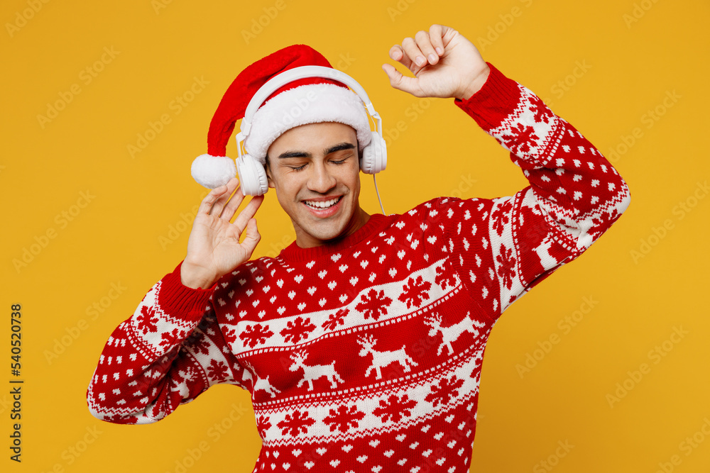 Merry young man wear red knitted christmas sweater Santa hat headphones posing listen music dance raise up hands isolated on plain yellow background. Happy New Year 2023 celebration holiday concept. Merry young man wear red knitted christmas sweater Santa hat headphones posing listen music dance raise up hands isolated on plain yellow background. Happy New Year 2023 celebration holiday concept.