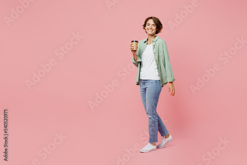 Full body side view young woman 20s wear green shirt white t-shirt hold takeaway delivery craft paper brown cup coffee to go isolated on plain pastel light pink background People lifestyle concept