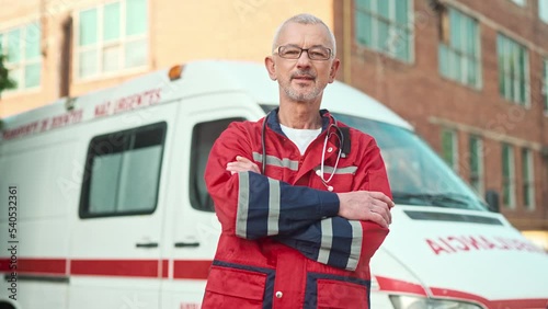 Camera approaching Caucasian middle-aged professional doctor in uniform standing outside near ambulance car and looking at camera. Male paramedic in glasses outdoor. Medical worker. First aid concept
