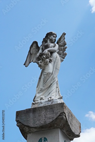 Vertical shot of a young female angel statue with spread wings holding flowers.  Female sculpted stone body with blue background.   