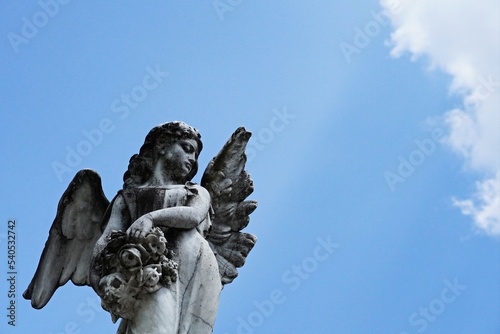 Young female angel sculpture with open wings isolated on blue sky background.  Antique sculpted angelic girl holding flowers and looking away from camera.