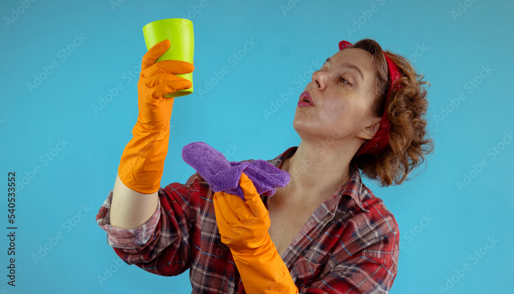 Pin-up woman in a plaid shirt and orange gloves wipes a green glass with a purple rag on an isolated background. Portrait of an adult woman cleaning. Woman looking at a glass