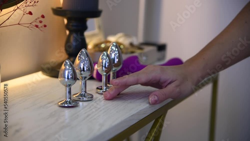 Close-up view of woman choosing sex toy and taking metal butt plug. Sex toys stands on white table. Real time video. Selective focus. Sexual issues theme. photo