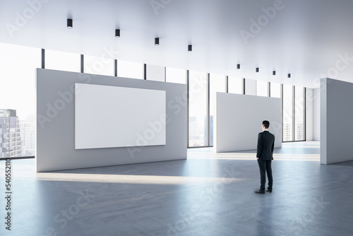 Businessman looking at blank white poster with space for your text or logo on light partitions in sunlit spacious exhibition hall with concrete floor and city view from panoramic windows, mock up