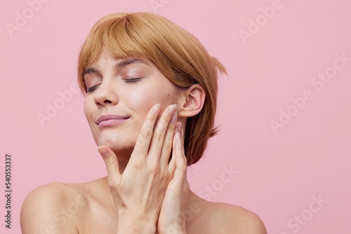 a close horizontal photo of a pleasant, lovely woman with perfect skin standing on a pink background gently touching her face with her hands, closing her eyes with pleasure