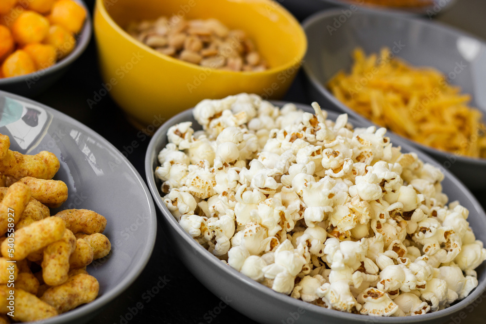 Salty snacks assortment shot on dark slate table. The picture includes potato chips, popcorn, peanut, cheese sticks, ketchup sticks, tzatziki chips, smoky flips, and others