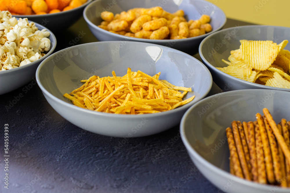 Salty snacks assortment shot on dark slate table. The picture includes potato chips, popcorn, peanut, cheese sticks, ketchup sticks, tzatziki chips, smoky flips, and others