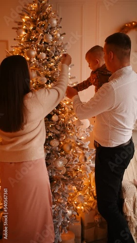 happy full-fledged joyful cheerful family man woman small child son in arms decorate Christmas tree toys garlands. heterosexual couple mom and dad parents celebrating new year together 4k vertical