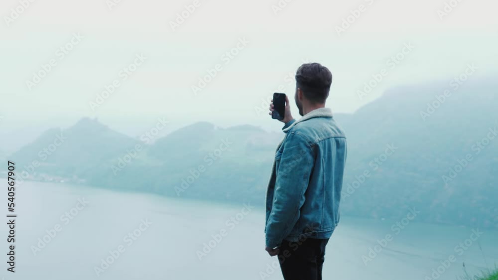 Young guy admiring swiss nature, mountains and the lake, standing and taking a photo on the cliff of a mountain. Happy excited man looking around at amazing scenery, foggy nature in autumn Young guy admiring swiss nature, mountains and the lake, standing and taking a photo on the cliff of a mountain. Happy excited man looking around at amazing scenery, foggy nature in autumn