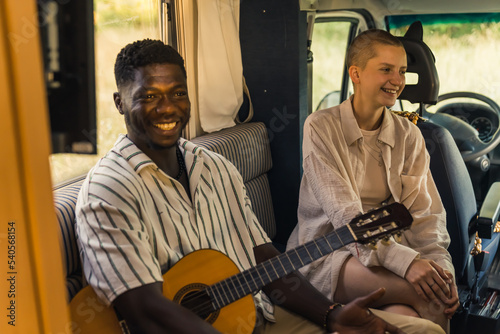 Indoor portrait of a couple of friends - handsome smiling black musician guy with acoustic guitar and friendly caucasian woman in white shirt - sitting on van sofa and laughing. High quality photo