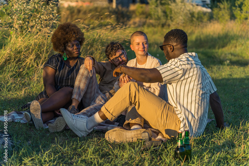 Outdoor party by the river. European youth. Multiethnic students in their mid 20s sitting on the grass, drinking beer, and talking. Meadow and golden hour sunshine. High quality photo