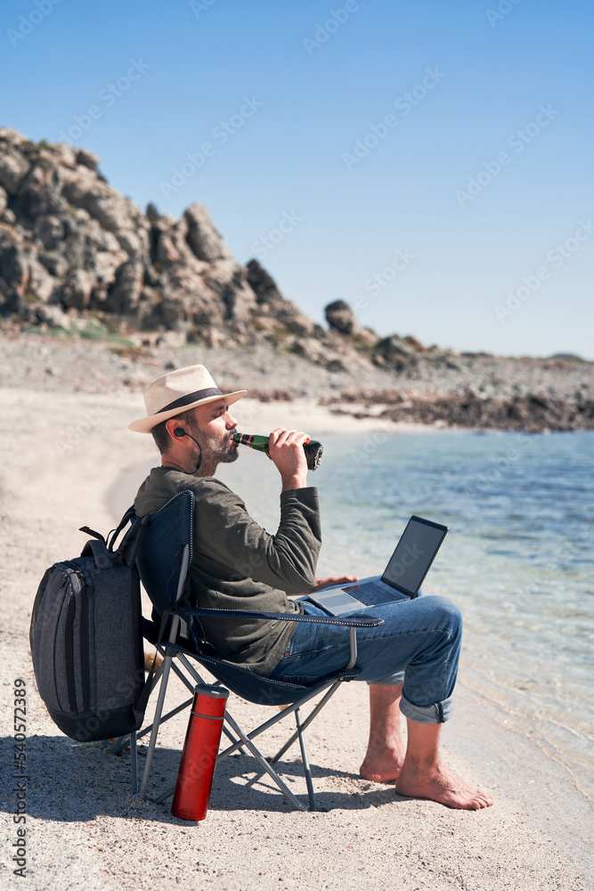 mature latin man sitting on the shore of the beach with a laptop and drinking beer and working