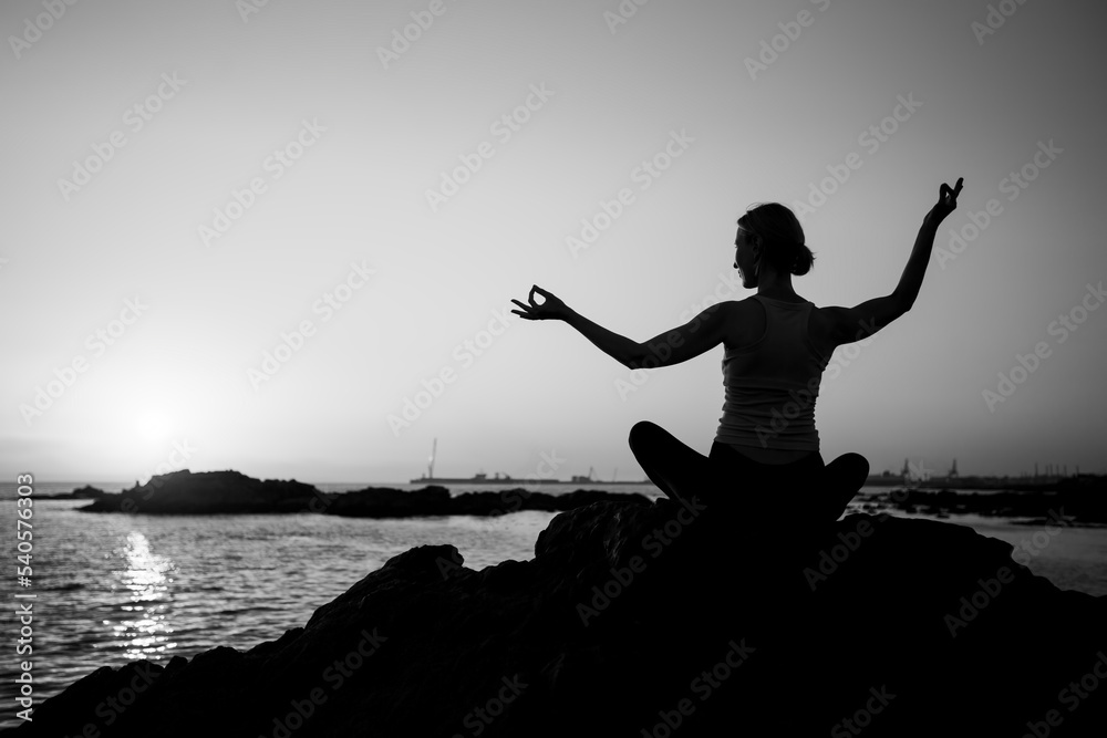 Silhouette of a woman practicing yoga during sunset on the Sea. Black and white photo.