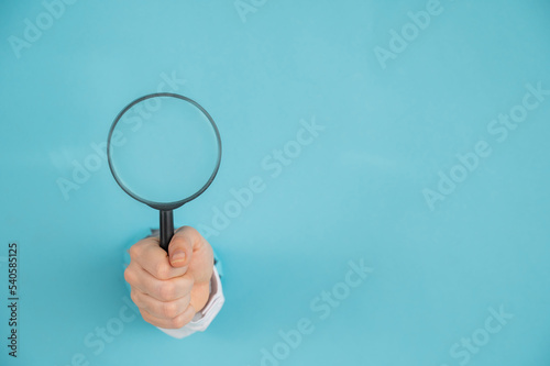 A woman's hand sticking out of a hole in a paper blue background holds a magnifying loupe. Copy space. 