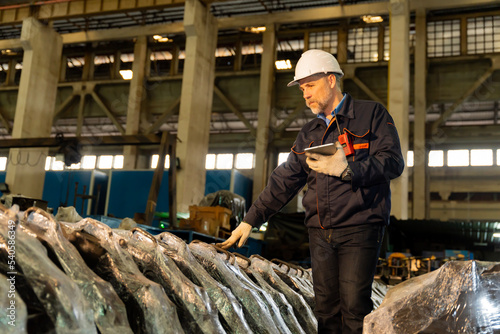 A machine factory engineer is inspecting the machines in the factory with a tablet or laptop and with a deliberate eye check. The factory is big and old.