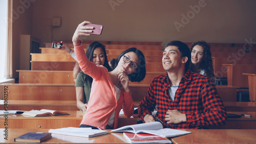Fotografia Cheerful students girls and guys are taking selfie in lecture hall sitting together at desks and holding smartphones