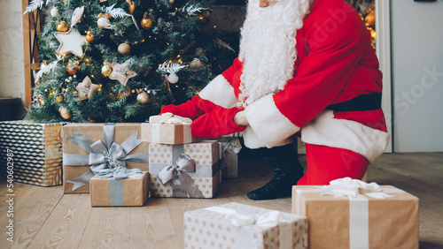 Santa Claus in traditional costume putting gift boxes under Christmas tree on holiday in house. Occasions and presents concept.