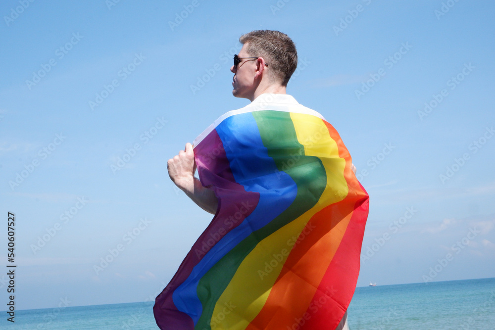 Young man holding a rainbow flag of LGBT symbols on a bright blue sky ...