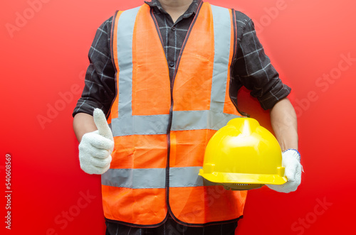 Front view of builder, engineer, consruction worker wear the orange safety vest and holding yellow construction helmet safety isolated over red background