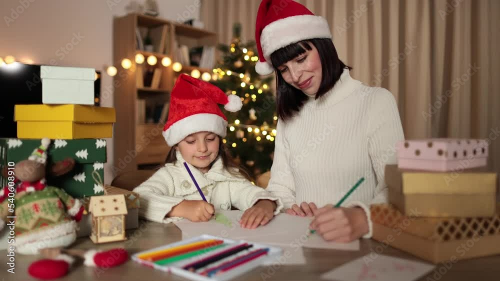 Preschool little girl wrapped up in writing letter to Santa Claus while sitting on table near her attractive mother with charming smile at decorated living room. Preschool little girl wrapped up in writing letter to Santa Claus while sitting on table near her attractive mother with charming smile at decorated living room.