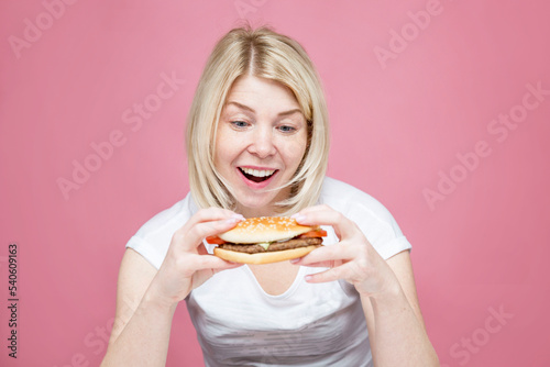 A young woman happily eats a hamburger. Beautiful blonde in a white shirt. Hunger  diets and junk delicious food. Pink background.
