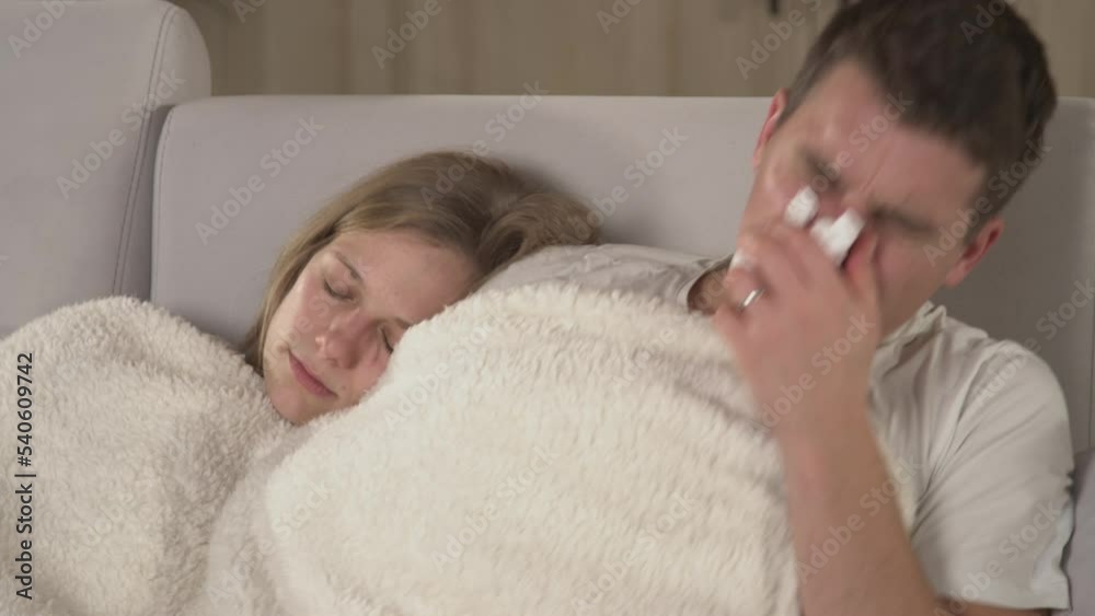 CLOSE UP: Young man sneezing and waking up his wife while they both rest on sofa. Married couple cuddling on comfortable couch while having a seasonal virus. Autumn flu and colds spreading around. CLOSE UP: Young man sneezing and waking up his wife while they both rest on sofa. Married couple cuddling on comfortable couch while having a seasonal virus. Autumn flu and colds spreading around.
