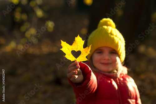 cheerful little girl in a red jacket and yellow hat holding a yellow maple autumn leaf with a heart  in the autumn park