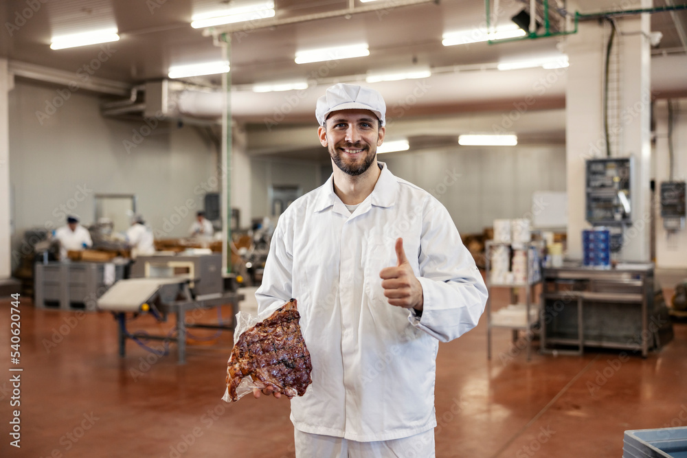 Butcher holds the peace of fresh meat and gives thumbs up while smiling at the camera and standing in the meat factory. Butcher holds the peace of fresh meat and gives thumbs up while smiling at the camera and standing in the meat factory.