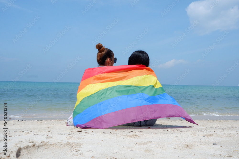 A lesbian couple holding a rainbow flag of LGBT symbols sits on the beach against a bright blue sky background. LGBT Concepts. A lesbian couple holding a rainbow flag of LGBT symbols sits on the beach against a bright blue sky background. LGBT Concepts.