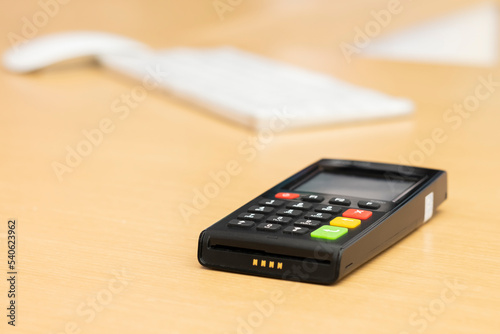 Black credit card machine POS-terminal (pin pad) modern device with NFC (near field communication) chip on wooden table. White keyboard and mouse as a bokeh background.