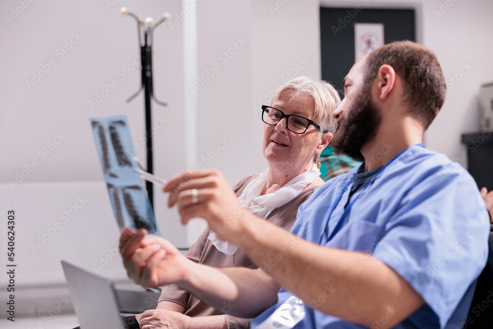 Obraz premium Male assistant hopsital staff showing x ray to elderly patient, in hospital reception lobby. Man nurse explaining radiography scan results to woman with disease, talking about treatment and diagnosis.