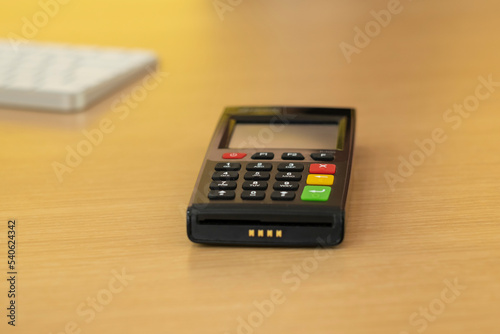 Black credit card machine POS-terminal (pin pad) modern device with NFC (near field communication) chip on wooden table. White keyboard as a bokeh background.