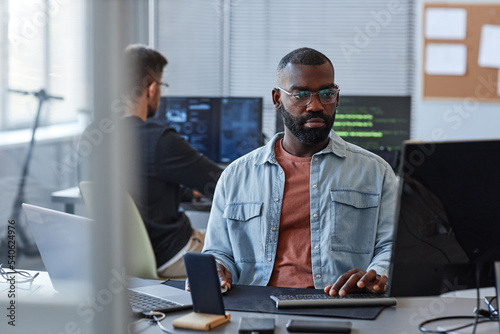 Portrait of black man using computer while programming mobile software in office behind glass wall