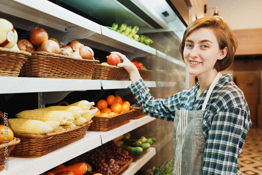 Portrait of Caucasian young adult saleswoman looking at camera with smile face and preparing to put an apple on the shelf. Female employee is adding apple to the stock of fruit in the grocery store.