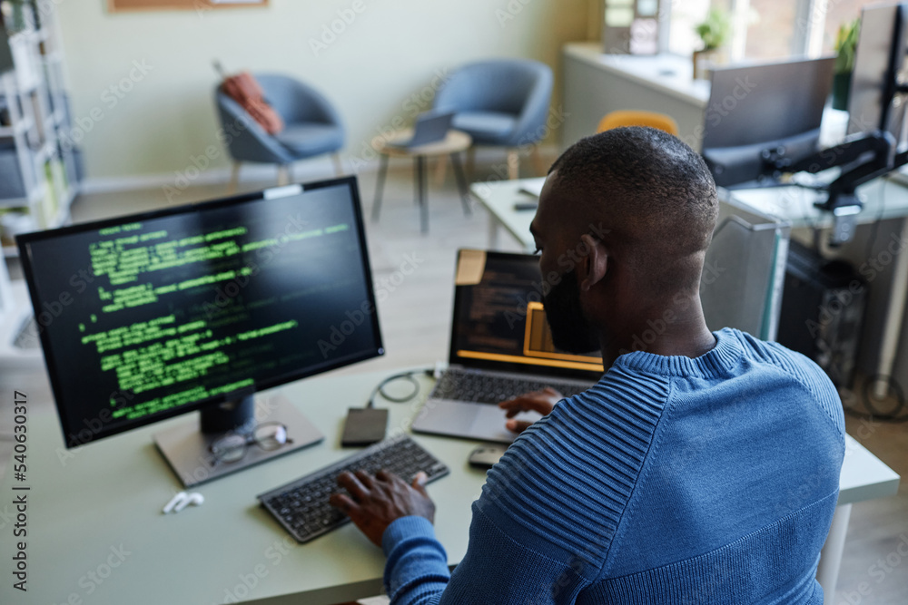 Back view of African American software developer writing code at workplace in office with multiple devices Back view of African American software developer writing code at workplace in office with multiple devices